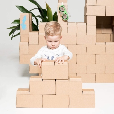 Criança a empilhar caixas de cartão marrons com planta e óculos verdes ao fundo