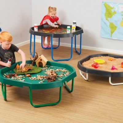 Três mesas de atividades infantis com diferentes materiais e cores, em sala luminosa com chão de madeira e mapa-múndi na parede