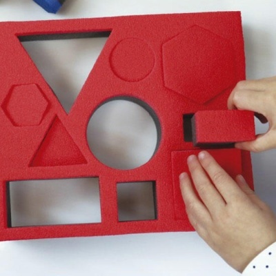 Painel educacional vermelho com formas geométricas e mãos a ajustar uma peça