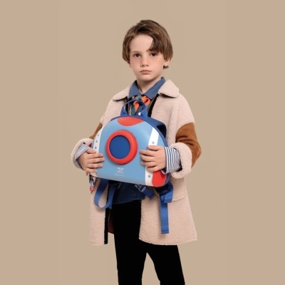 Criança segurando mochila infantil azul, vermelha e cinza, vestindo casaco bege com detalhes castanhos