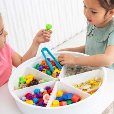 Crianças a brincar com uma tábua circular branca dividida em seis compartimentos com materiais coloridos e texturizados.