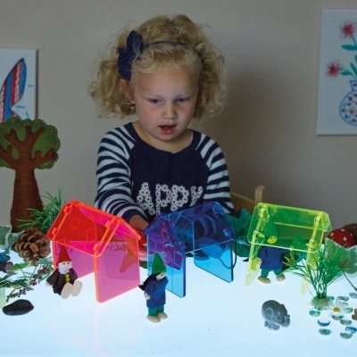 Menina loira a brincar com casinhas coloridas de plástico num ambiente com plantas e figuras decorativas.
