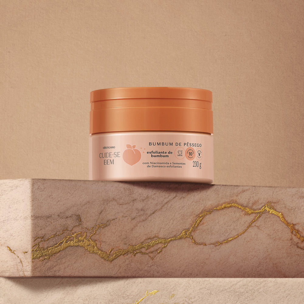 Creme esfoliante para corpo BUM BUM DE PÊSSEGO em embalagem redonda cor de pêssego com tampa terracota sobre superfície mármore bege com detalhes dourados
