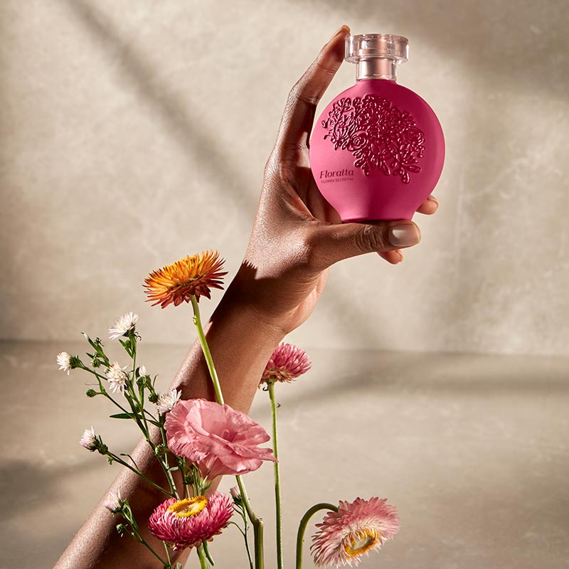 Floratta Flores Secretas Eau de Toillete 75ml Frasco de perfume rosa com relevo e tampa transparente segurado por mão junto a flores naturais