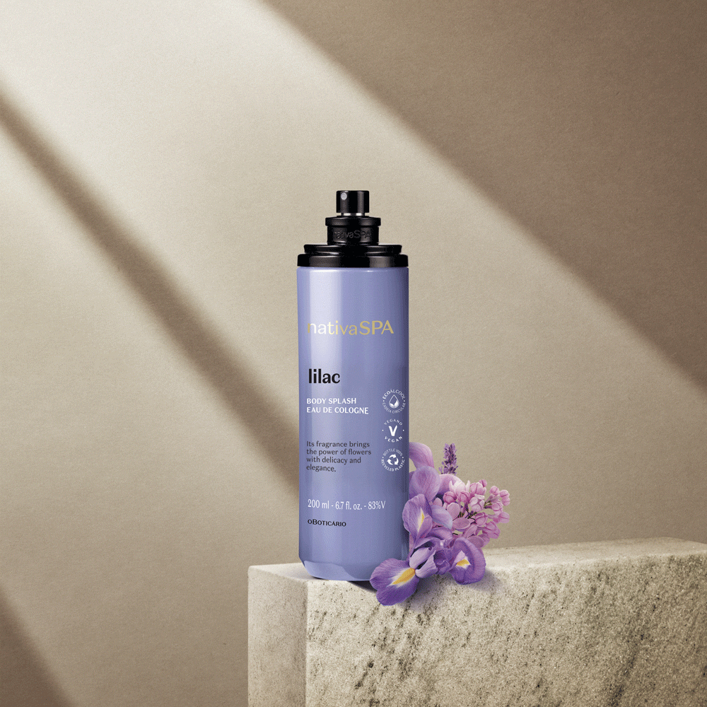 Frasco lilás de body splash com flores lilases ao lado sobre superfície cinza