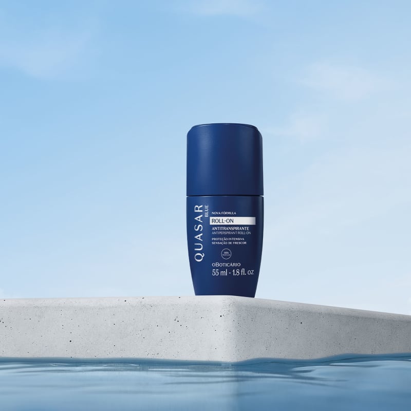 Quasar Blue Desodorante Roll-On 55ml Frasco azul escuro de roll-on antitranspirante Quasar sobre superfície de cimento com céu azul e água.