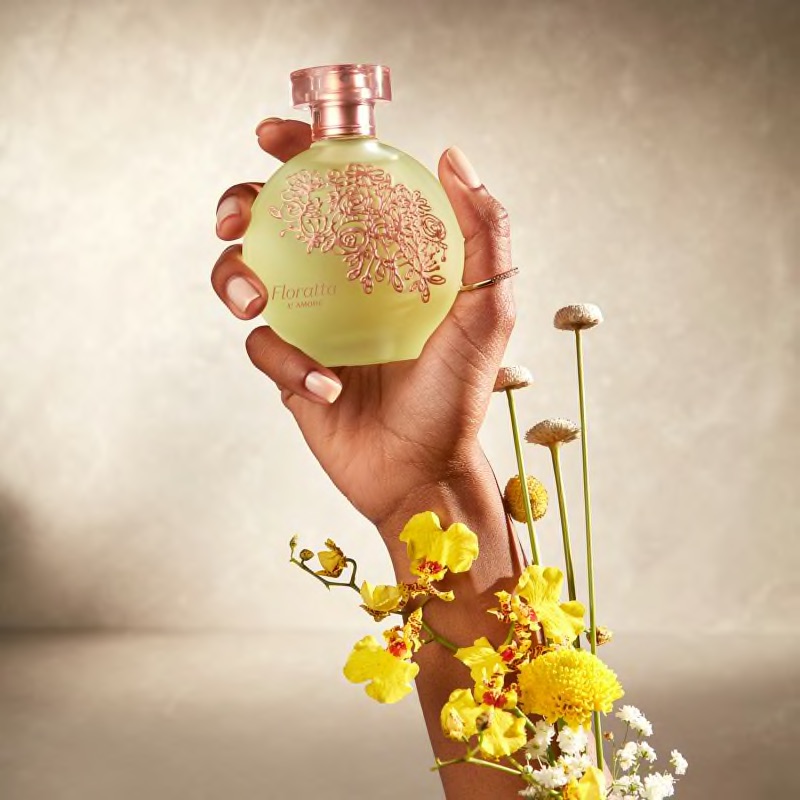 Floratta L'Amore Eau de Toilette 75ml Frasco redondo de perfume amarelo com tampa rosa e padrão floral segurado por mão com flores amarelas.