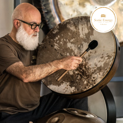 Homem idoso toca tambor redondo com baqueta em ambiente com tambores