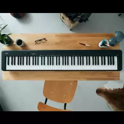 Piano digital preto com 61 teclas sobre mesa de madeira com vários objetos ao redor
