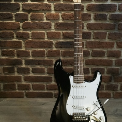 Guitarra elétrica preta com escudo branco e braço de madeira numa parede de tijolos