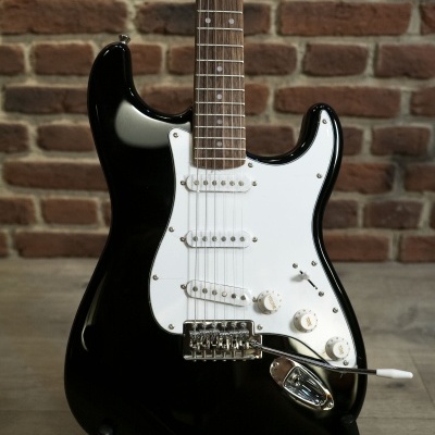 Guitarra elétrica preta com escudo branco e três captadores sobre suporte numa sala com parede de tijolos.