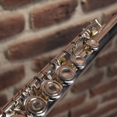 Flauta transversa metálica prateada com teclas circulares e detalhes dourados com parede de tijolos ao fundo