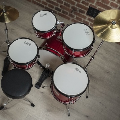 Conjunto de bateria vermelha com tambores e pratos montados em suporte, banco preto, piso de madeira e parede de tijolos