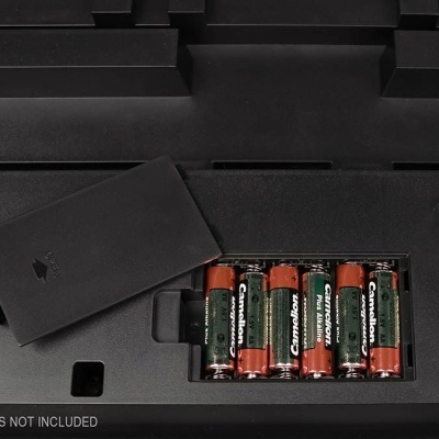 Compartimento de pilhas aberto em dispositivo preto com pilhas AA visíveis e texto BATTERIES NOT INCLUDED.