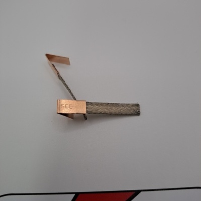 Peça metálica com lâminas de cobre e faixa cinzenta com inscrição SCB sobre fundo cinzento claro e logótipo vermelho e preto