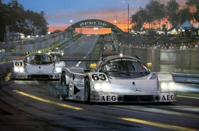 Dois carros de corrida Mercedes-Benz prateados na pista com arco Dunlop ao fundo