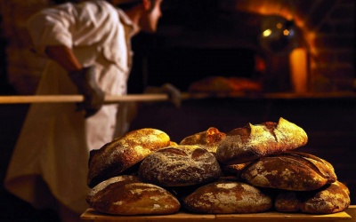 Pães artesanais empilhados em madeira com padeiro ao fundo numa padaria