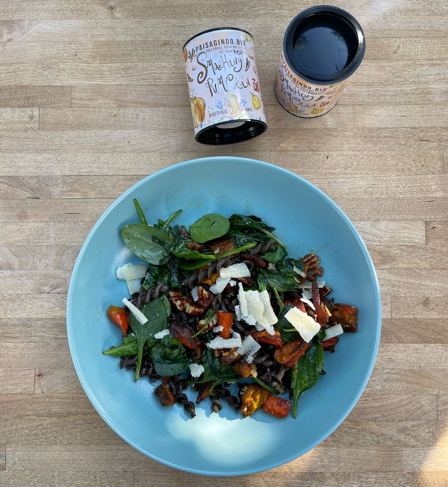 Tigela azul com salada colorida e dois frascos redondos de tempero numa mesa de madeira.