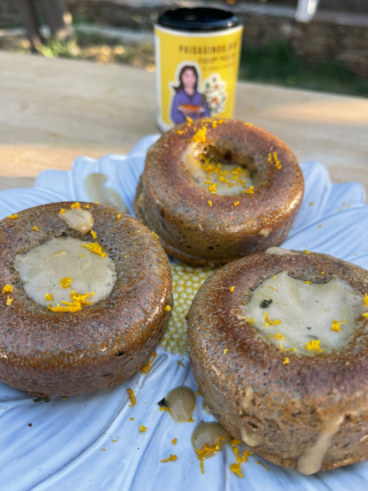 Muffin masala com creme de coco e laranja Três bolinhos castanhos com cobertura cremosa e flocos amarelos num prato branco, frasco amarelo ao fundo.