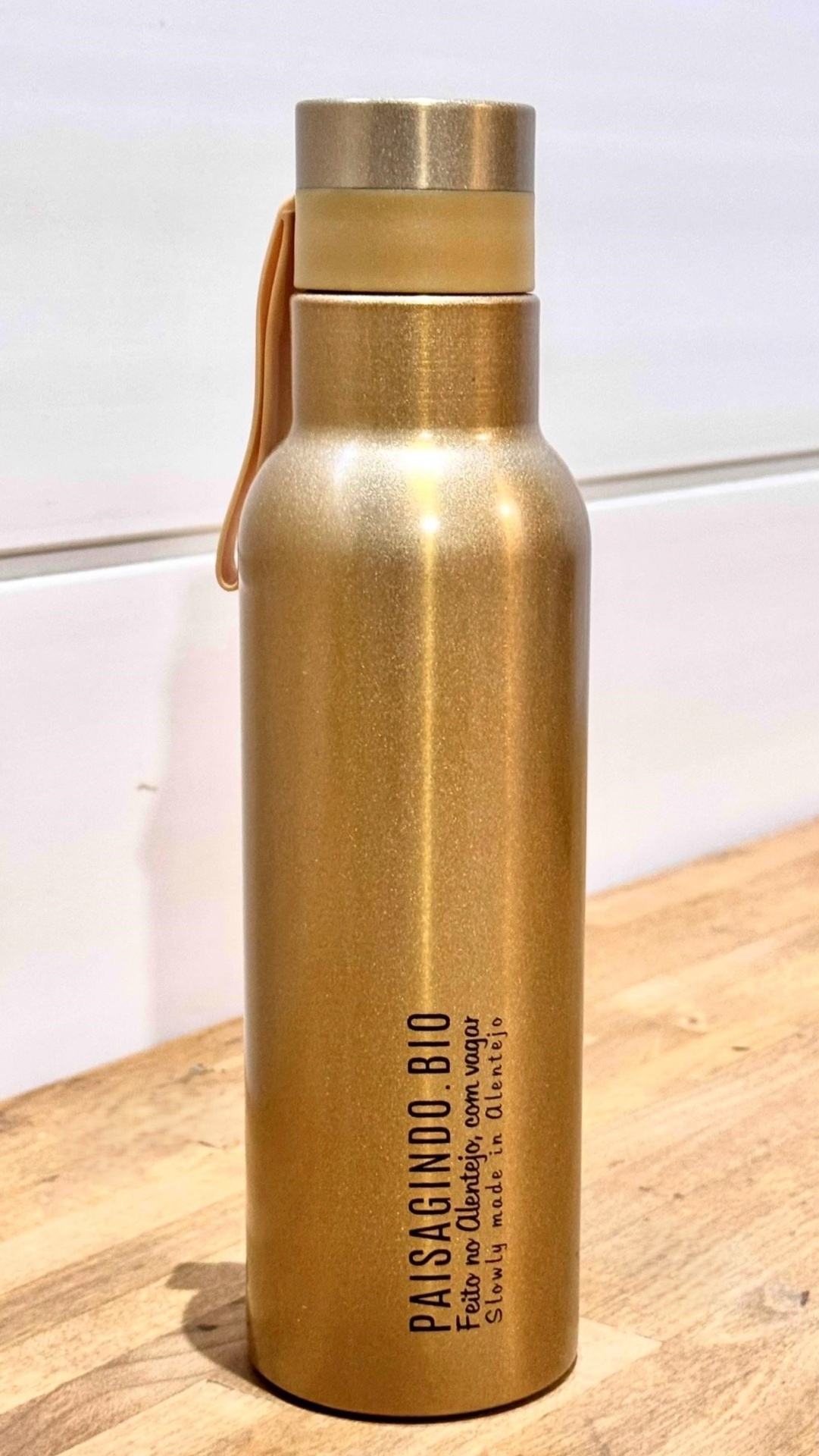 Garrafa térmica "dourada" em inox (530 ml) Garrafa metálica dourada com texto preto 'PAISAGINDO BIO' e alça de silicone laranja