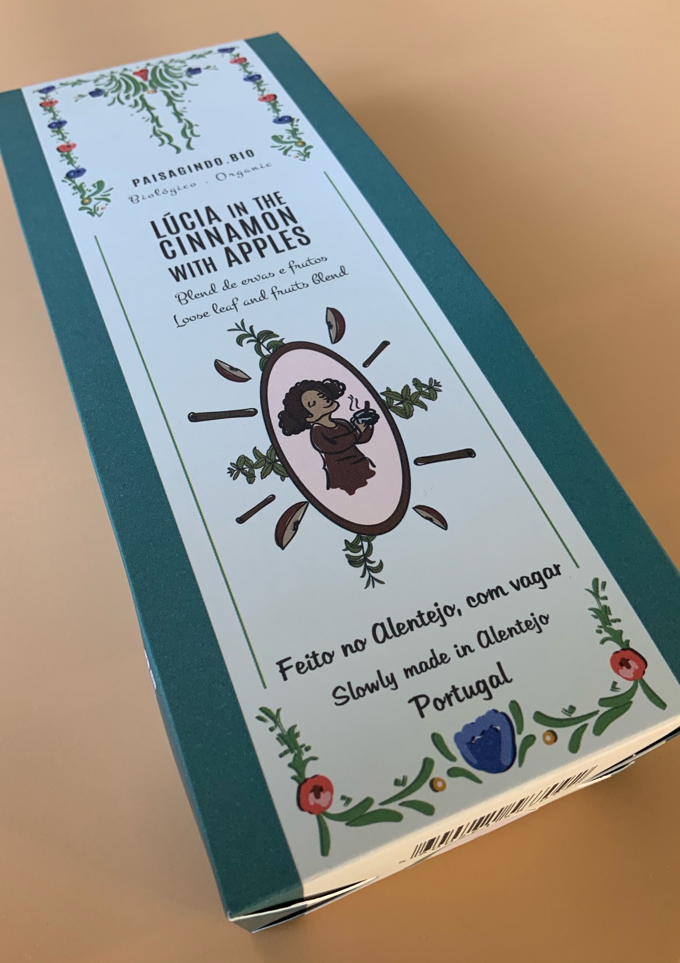 Lúcia in the Cinnamon with Apples (Apple & Cinnamon) Embalagem de produto alimentar com textos em inglês e português, ilustração de mulher e decoração floral.
