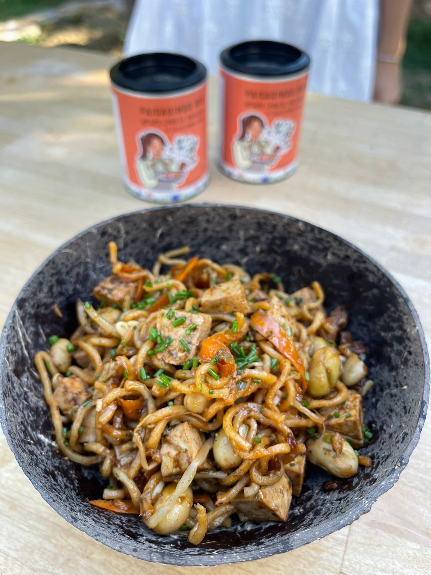 Prato de noodles com tofu e legumes num prato preto, duas latas alaranjadas ao fundo numa mesa de madeira.