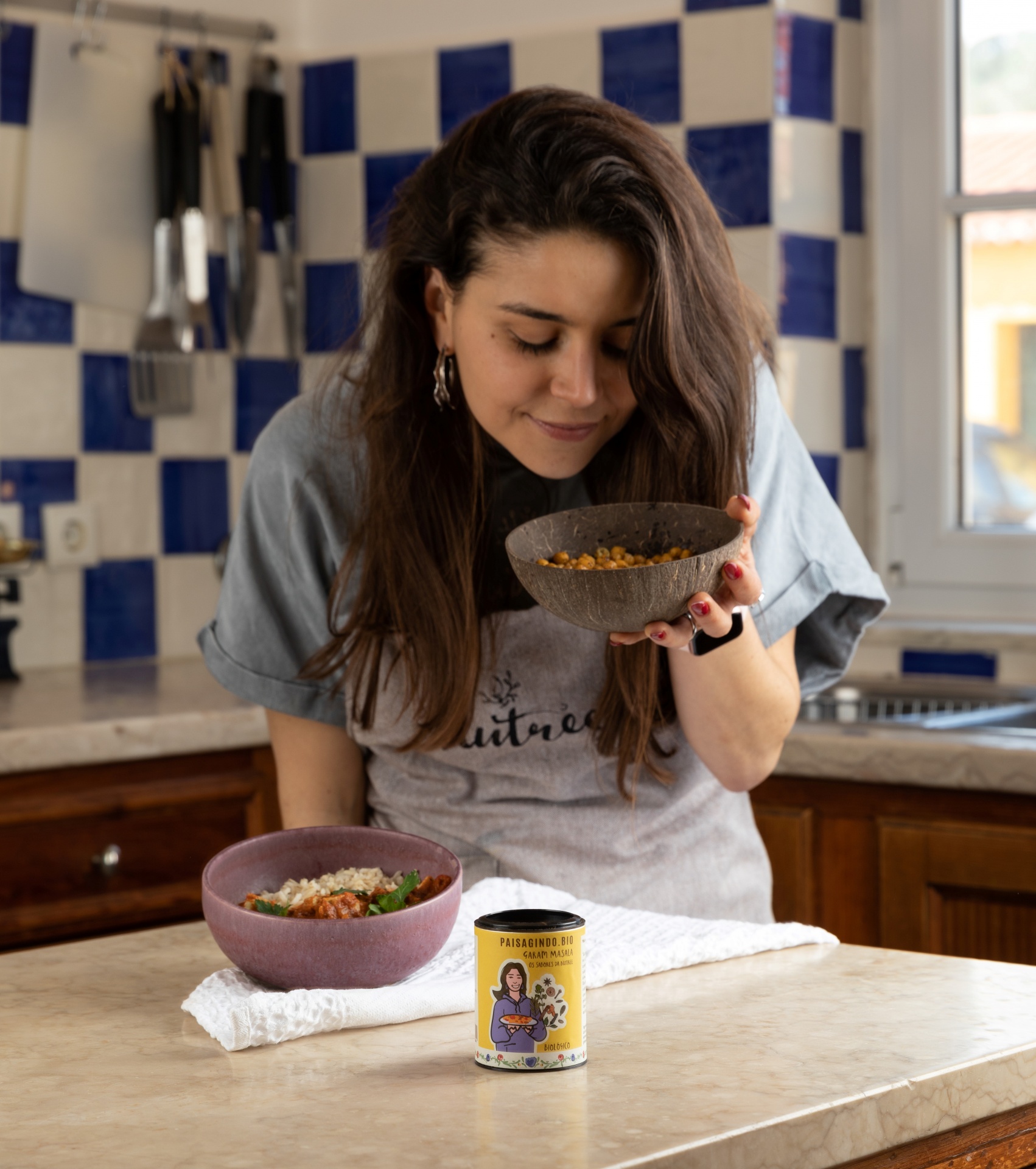 Garam Masala by Os Sabores da Nutree Frasco amarelo de pasta de amendoim e tigelas roxas com comida numa bancada de cozinha com azulejos azuis e brancos.