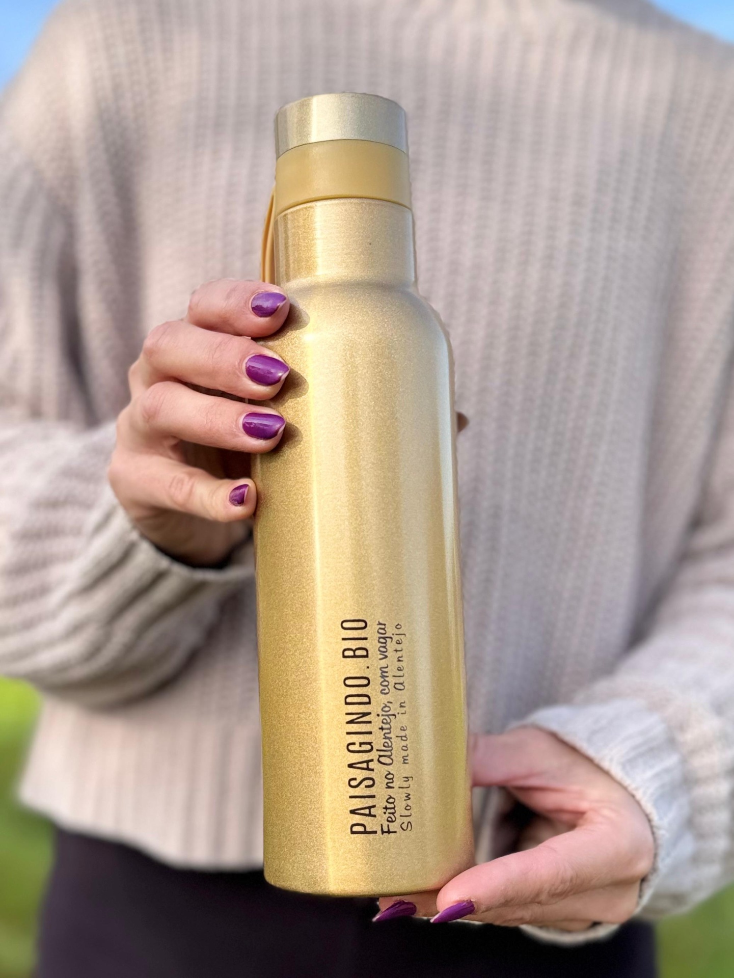 Garrafa térmica "dourada" em inox (530 ml) Garrafa metálica dourada com texto PAISALGINO BIO e tampa, segurada por mãos com unhas roxas e camisola de malha bege.