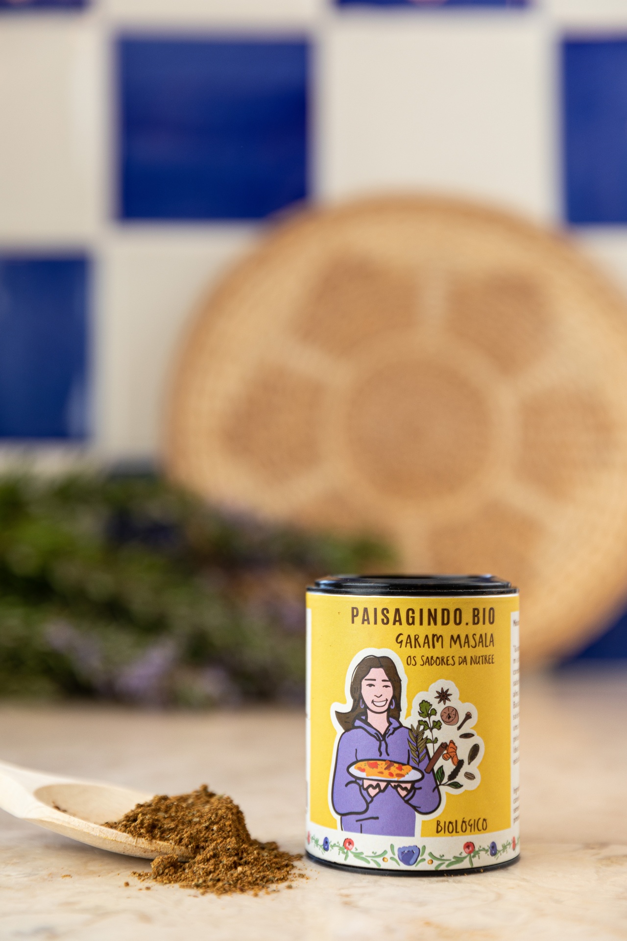 Garam Masala by Os Sabores da Nutree Frasco de especiarias Garam Masala biológico amarelo com colher de madeira e pó castanho sobre bancada com azulejos azuis no fundo