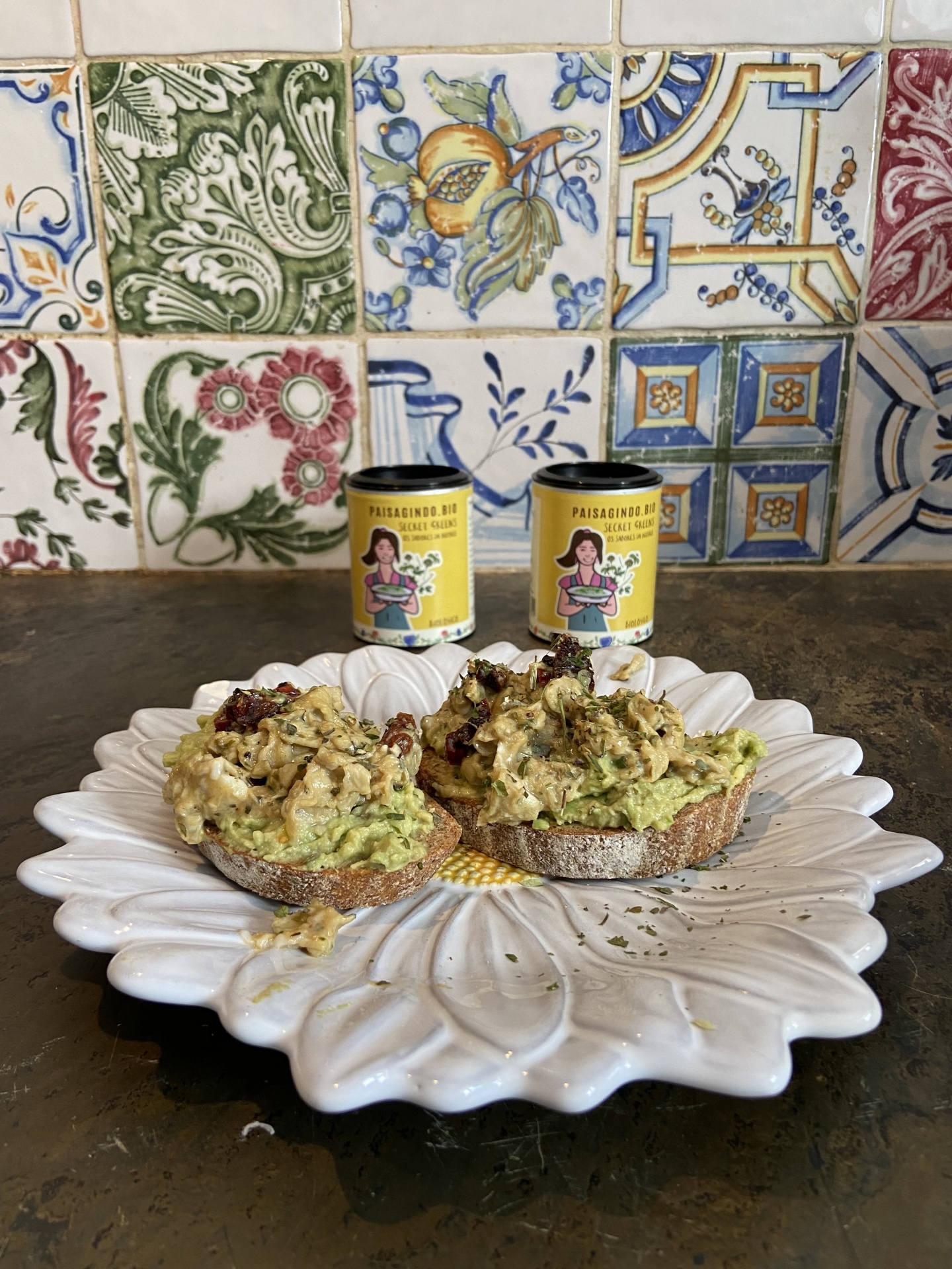 Eggs Toast & Secret Greens Tostas com pasta verde em prato branco e dois frascos amarelos em fundo de azulejos coloridos