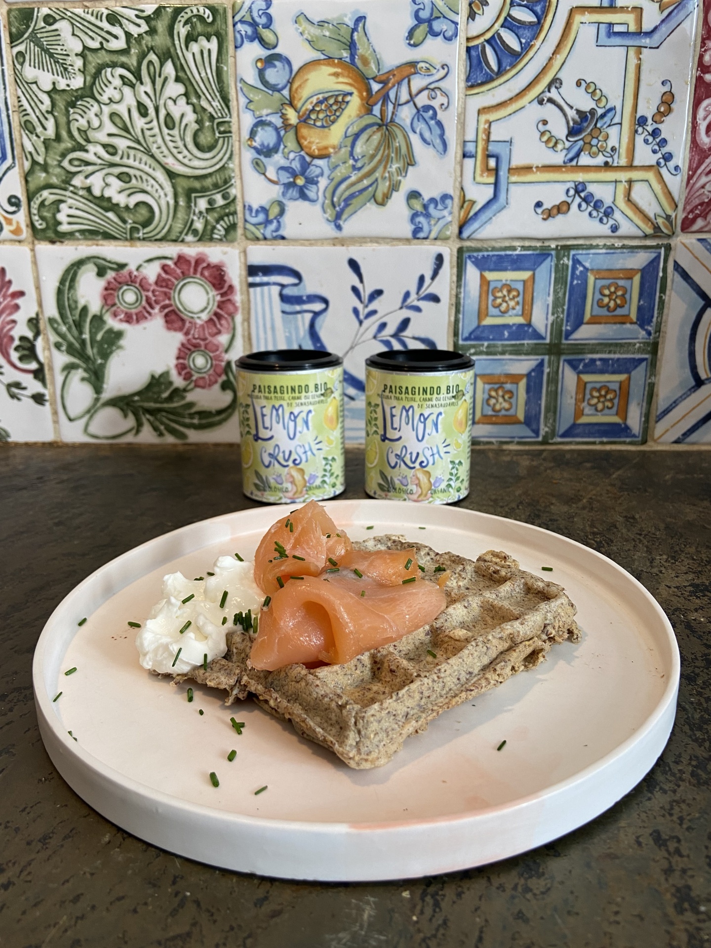 Waffle salgado com Lemon Crush Prato branco com waffle multigrãos, salmão fumado, creme branco e cebolinho, diante de azulejos coloridos e latas de tempero amarelas.