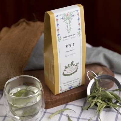 Pacote de stévia bio com infusão em copo de vidro e folhas secas sobre pano xadrez