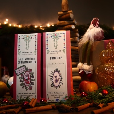 Duas caixas de chá natalícias decoradas, folhas verdes, paus de canela, pinhas e decoração festiva em fundo iluminado.