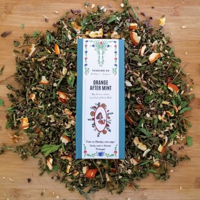 Pacote branco de chá Orange After Mint rodeado por folhas e pedaços secos de chá sobre mesa de madeira