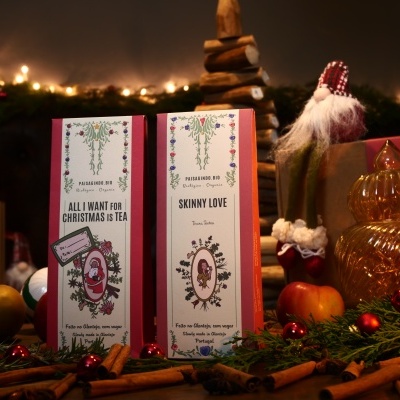 Dois pacotes de chá decorativos com texto em inglês rodeados de decoração natalícia incluindo paus de canela e maçãs