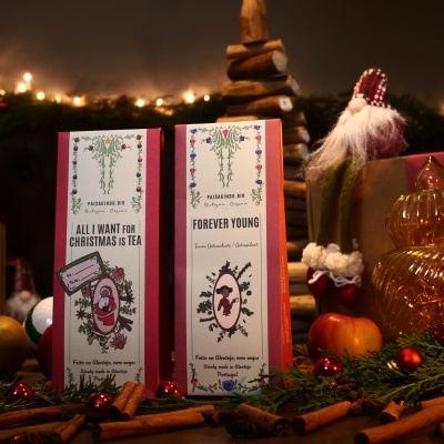 Dois pacotes de chá festivos com texto em inglês e decoração natalícia sobre uma mesa com canela e folhagens verdes