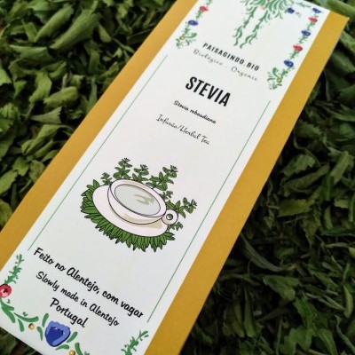 Caixa amarela de chá de Stevia com rótulo branco decorado, sobre folhas verdes secas.