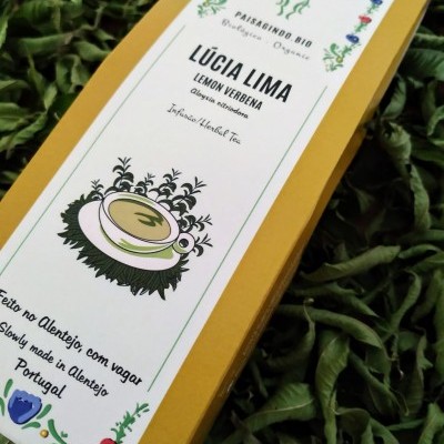 Embalagem amarela com rótulo branco de infusão herbal orgânica de lúcia-lima sobre folhas verdes.