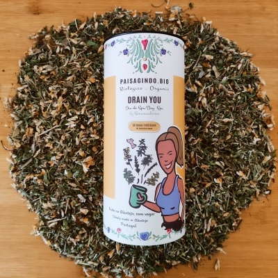 Embalagem cilíndrica branca de chá orgânico Drain You rodeada por ervas secas sobre madeira