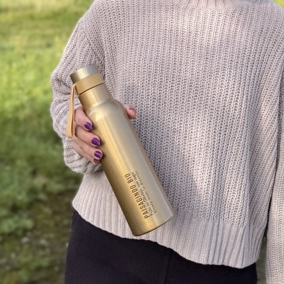 Garrafa térmica dourada segurada por pessoa com casaco de malha cinza em exterior com relva