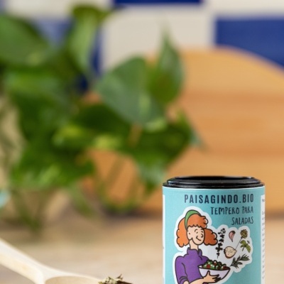 Frasco de tempero para saladas biológico PAISAGINDO.BIO com ilustração colorida ao lado de ervas numa colher de madeira