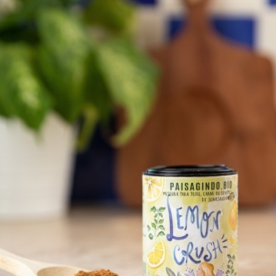 Embalagem cilíndrica com rótulo colorido 'LEMON CRUSH' junto a colher com pó castanho, sobre bancada, planta e tábua ao fundo