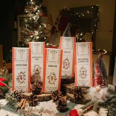 Cinco caixas decorativas brancas com laranja e texto natalício, rodeadas por elementos naturais e decoração de Natal com árvore iluminada ao fundo