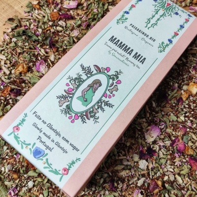 Caixa rosa com rótulo branco e ilustração de mulher, rodeada por ervas secas e flores sobre madeira