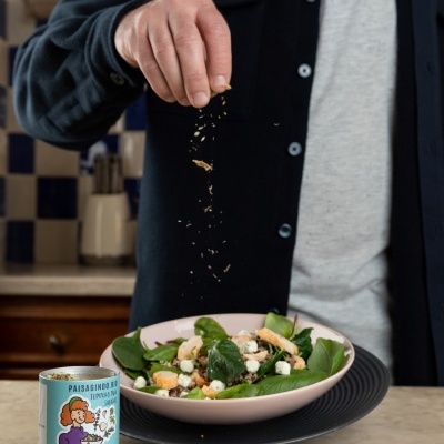 Pessoa polvilhando tempero numa salada com embalagem PAISAGINO II TEMPEROS AO LAR ao lado