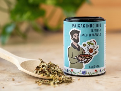 Embalagem azul clara de tempero mediterrânico biológico com ilustração e texto, sobre superfície de madeira com colher e ervas secas