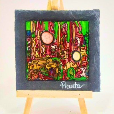 Quadro pequeno colorido com dois relógios embutidos em moldura de pedra sobre cavalete de madeira.