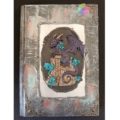 Livro com capa cinzenta texturizada e dragão tridimensional num castelo com flores azuis