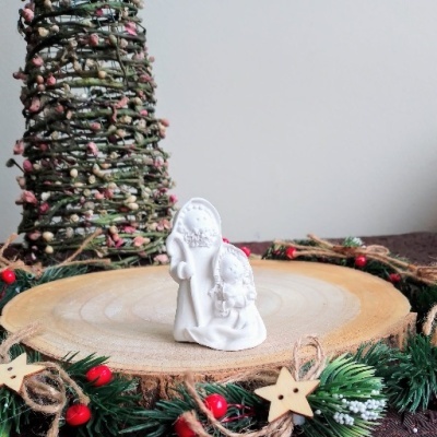 Figura decorativa natalícia branca de São José e Menino Jesus sobre disco de madeira com arranjo verde e bagas vermelhas