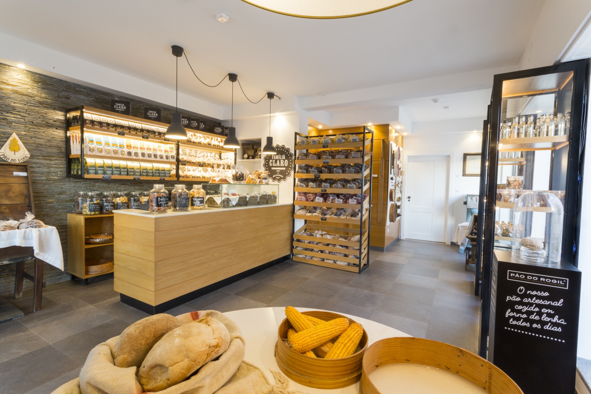 Interior de loja com balcão em madeira, produtos em prateleiras, pães e milho em cestos de madeira